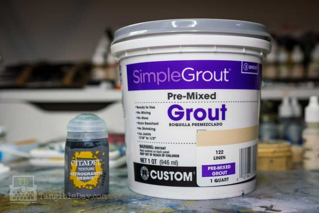Side-by-side size comparison of a small Citadel Texture Paint pot next to a one-quart container of premixed sanded tile grout used for miniature basing
