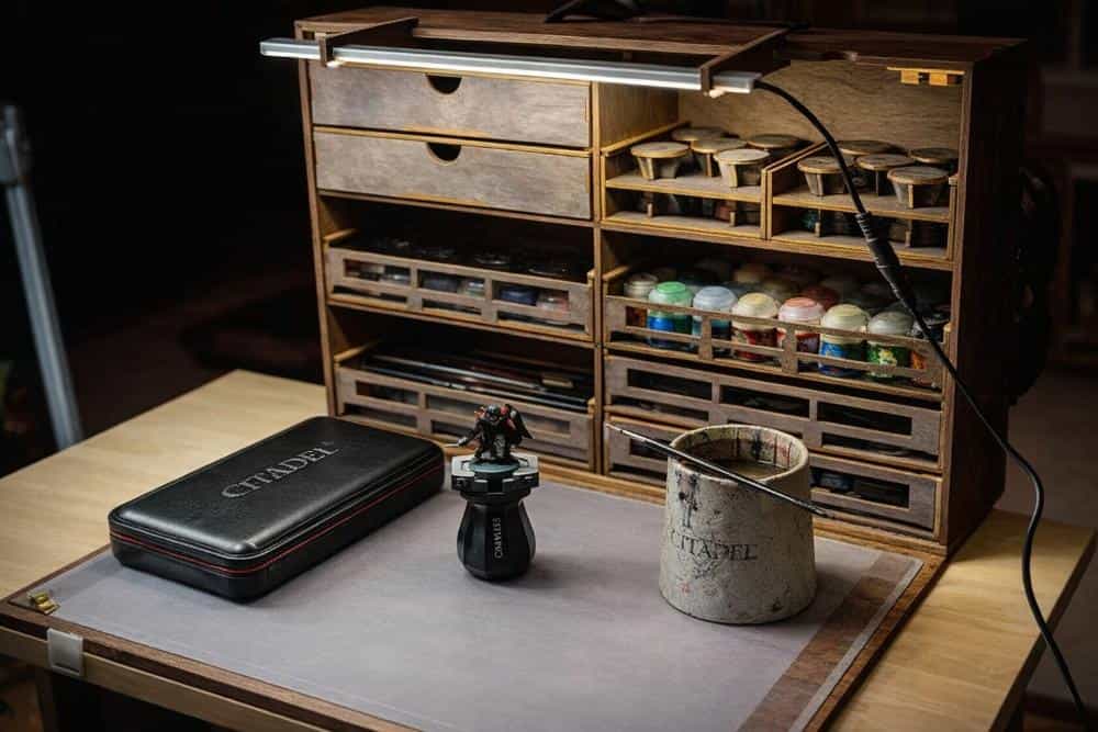 Portable hobby workstation with organized miniature paints, drawers, brush holders, and LED lighting on a wooden painting desk