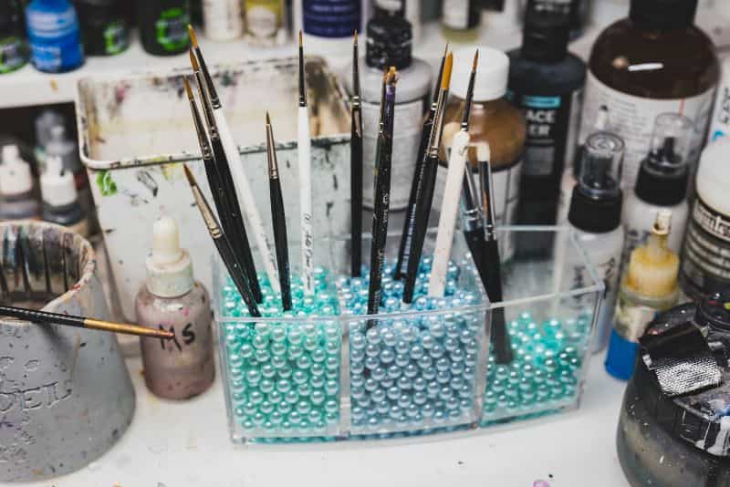 Miniature painting brushes organized upright in container filled with beads on hobby desk