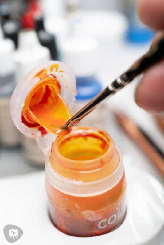 Close-up of a miniature painting brush dipping into a pot of bright orange and yellow paint