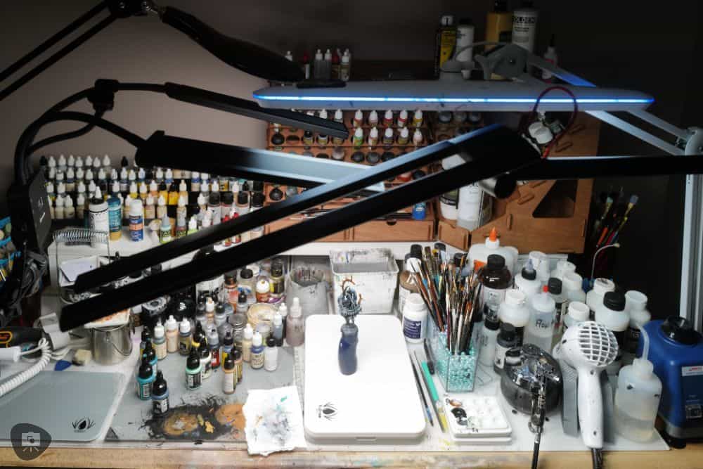 Busy hobby desk setup with three articulated task lamps and paint supplies