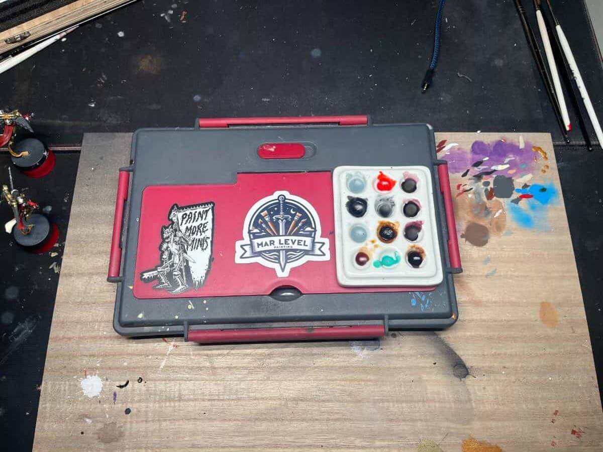 A portable painter’s wet palette with lid with multiple paint wells, some filled with various colors, on a wood-stained table. 
