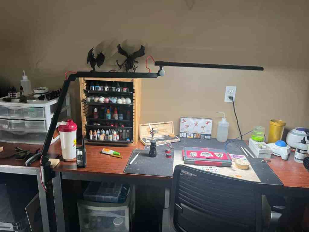 A full view of a home workstation lit by the Redgrass R9 desk lamp, showing a spread of hobby tools and paints