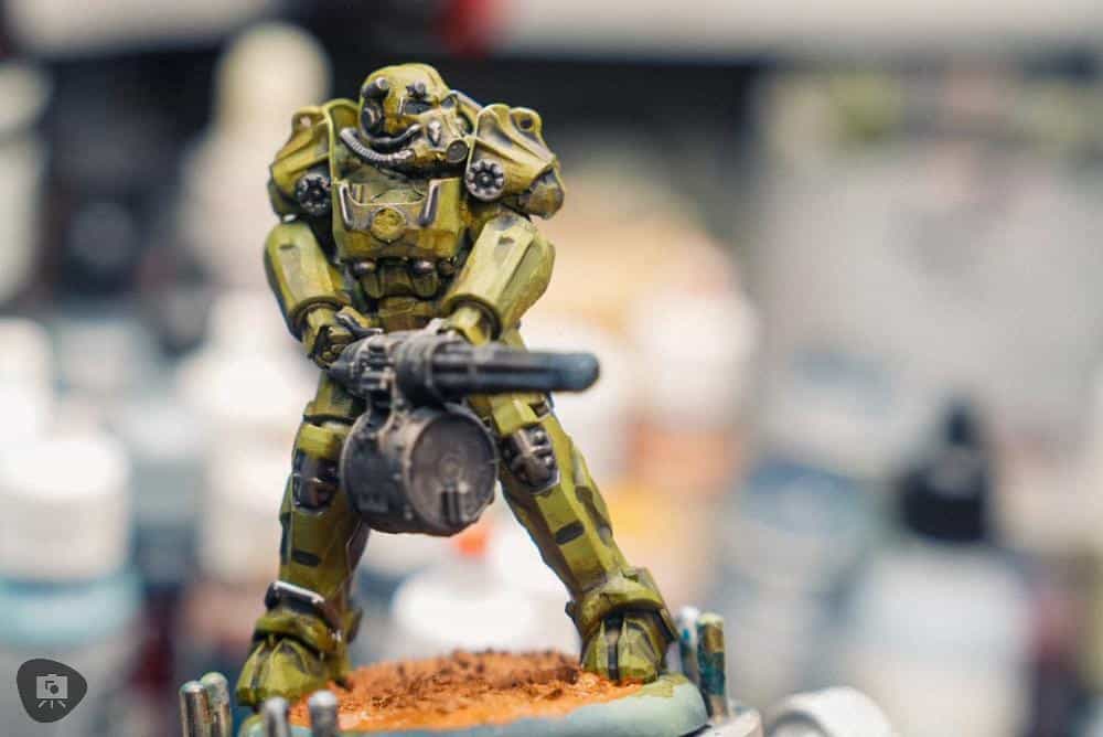 A painted Fallout miniature in power armor, posed with a minigun, against a blurred hobby workspace background.