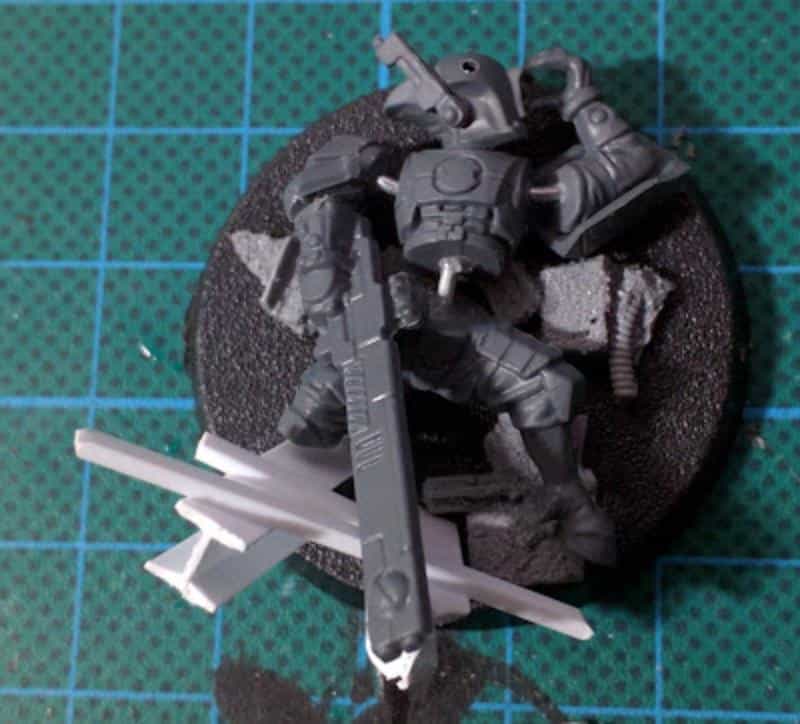 Sci-fi miniature standing on a textured base with white sprue shards arranged as battlefield debris, shown on a green cutting mat during assembly.