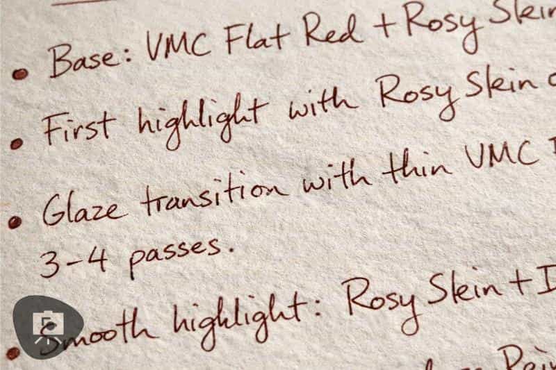 Close-up of handwritten notes in red ink on a notebook page, showing a rough miniature painting recipe with short bullet points and glazing steps.